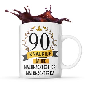Könnte beinhalten: Weiße Keramik-Kaffeetasse mit einem schwarzen und goldenen Design. Die Tasse zeigt die Zahl 90, die Worte "Knackige Jahre" und den Spruch "Mal knackt es hier, mal knackt es da". Kaffee spritzt in die Tasse.