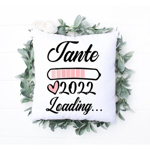 Könnte beinhalten: Weißes quadratisches Kissen mit dem Wort "Tante" in schwarzer Schrift, einer Ladebalken-Grafik, dem Jahr "2022" und dem Wort "Loading..." in Schwarz. Das Kissen ist von grünem Laub und weißen Blumen umgeben.