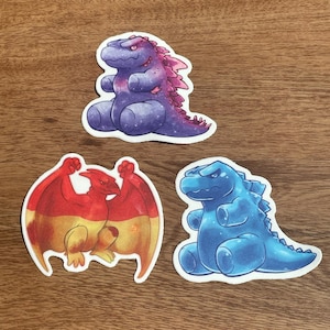 Puede incluir: Tres pegatinas coloridas con dinosaurios de dibujos animados. Una es morada con detalles rosas, otra es roja y amarilla con alas, y la última es azul. Las pegatinas están sobre una superficie de madera.