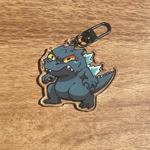 以下が含まれることがあります： 象徴的な怪獣の漫画イラストが特徴の、ゴジラをテーマにしたキーホルダー。キーホルダーは黒い留め金と透明なアクリルボディを備えています。ゴジラのフィギュアは、黄色い目と白い背びれを持つ濃い青色です。