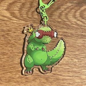 May include: A green Tyrannosaurus Rex keychain with a gold crown and a red and green snack in its mouth. The keychain has a green clasp and is made of clear acrylic.