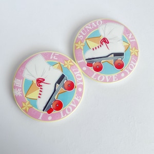 May include: Two round, pink and blue stickers with a white roller skate design and the text "SUNAO NI LOVE YOU" in a circle. The roller skates have red wheels and laces. The background includes stars.