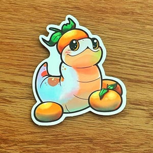 May include: A colorful sticker featuring a cartoon caterpillar with an orange hat and two oranges. The caterpillar has a light blue and orange body with a white outline. The oranges are orange with green leaves.