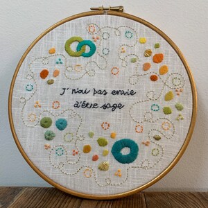 Peut inclure: Broderie sur toile dans un cercle en bois doré. Le texte en français « J'ai pas envie d'être sage » est entouré de cercles et de motifs brodés en teal, orange, vert et jaune. Le fond est en lin.