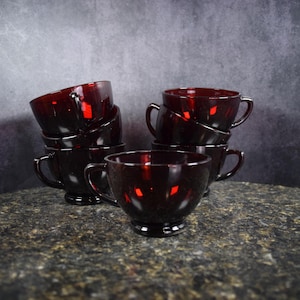 May include: A set of eight ruby red glass cups with handles. The cups are stacked in two piles, with one pile of four cups and another pile of four cups. The cups are on a dark gray granite countertop.