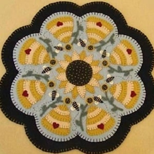 May include: A circular, blue and yellow felt rug with a sunflower design in the center. The sunflower has a black center with white dots and yellow petals. The rug is surrounded by a black border with a scalloped edge.