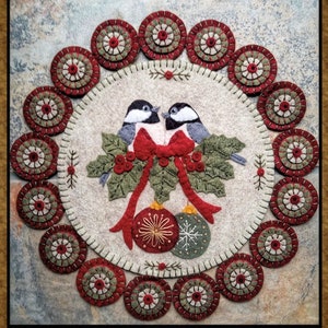 May include: A felt Christmas ornament with a red and green holly design, two birds, and a red bow. The ornament is surrounded by a circle of smaller felt ornaments.