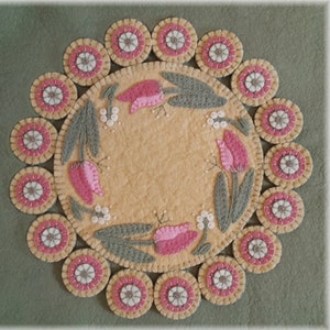 May include: A round, cream-colored felt rug with a border of pink and white circles. The center of the rug features a design of pink tulips with green leaves and white beads.