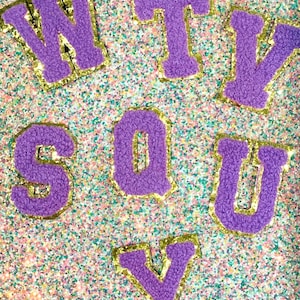 May include: Purple felt letters spelling out "SQUY" with a gold outline on a glittery background.