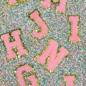 May include: Pink, fluffy varsity-style letter patches with gold glitter edging are scattered across a glittery background. The letters include K, M, J, I, H, N, G, and L. These patches are likely for customising apparel or accessories.