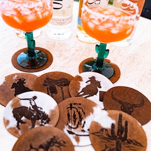 Peut inclure: Ensemble de dessous de verre ronds avec des motifs sur le th&egrave;me de l'Ouest, dont des cowboys, des selles et des cactus. Les dessous de verre sont en peau de vache marron et blanche. Deux cocktails et des bouteilles de tequila sont visibles.