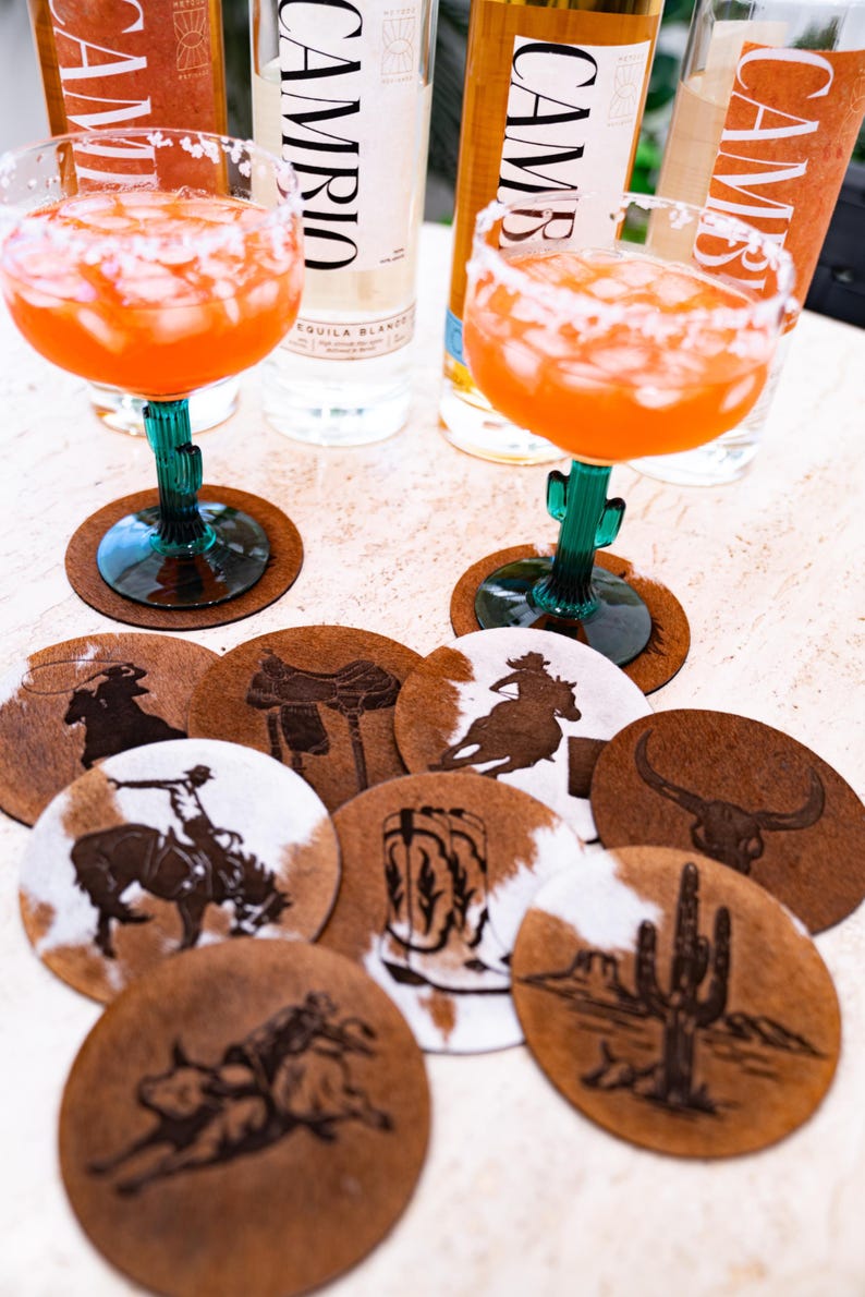 Peut inclure: Ensemble de dessous de verre ronds avec des motifs sur le th&egrave;me de l'Ouest, dont des cowboys, des selles et des cactus. Les dessous de verre sont en peau de vache marron et blanche. Deux cocktails et des bouteilles de tequila sont visibles.