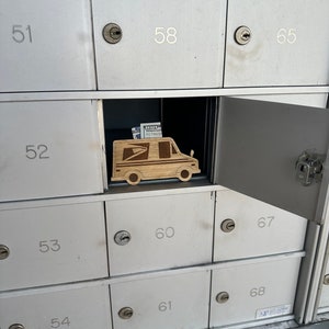May include: A wooden USPS mail truck is inside an open mailbox. The mailbox is one of many in a bank of silver mailboxes. The mail truck is made of light-colored wood and has the USPS logo. There is a stack of US $100 bills inside the mail truck.