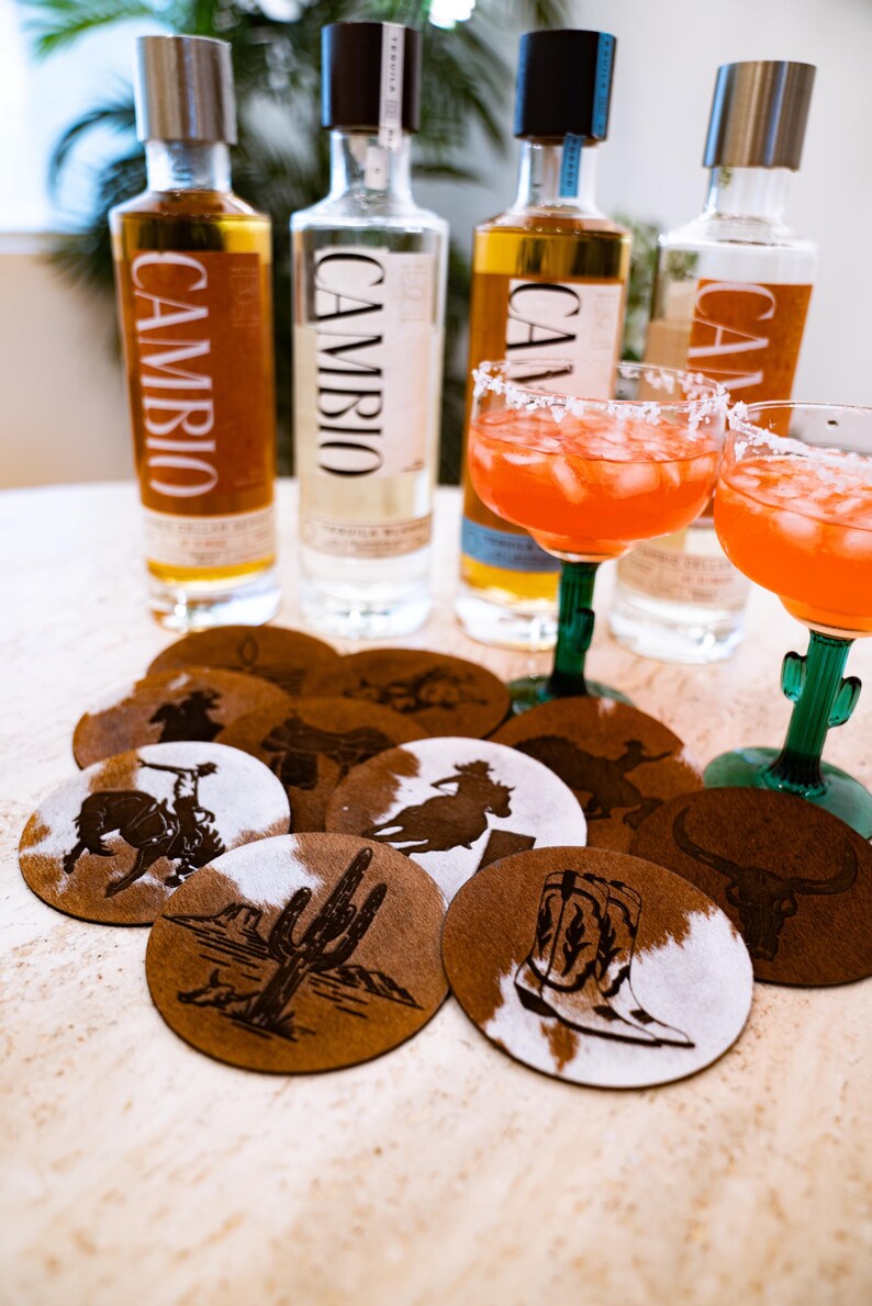 Peut inclure: Nature morte avec des bouteilles de tequila, des cocktails et des dessous de verre. Les bouteilles de tequila portent l'inscription "CAMBIO". Les dessous de verre ont un motif en peau de vache et des motifs sur le th&egrave;me de l'Ouest.