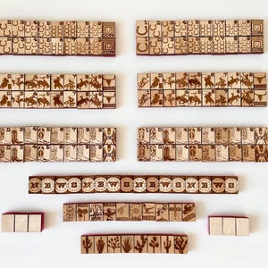 May include: Wooden game tiles with engraved Western-themed designs, including horseshoes, cowboys, boots, and cacti. The tiles are arranged in rows and columns, with some featuring the word "NEWS". The tiles are light brown with dark brown engravings.