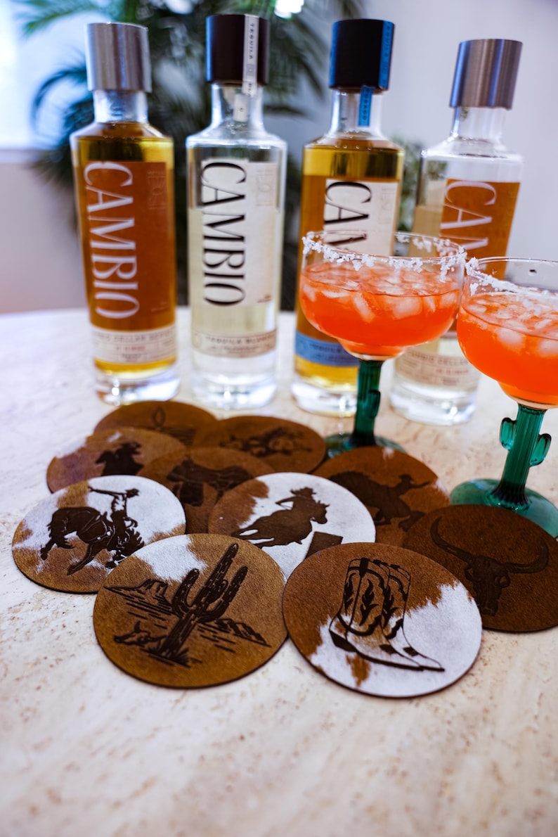 Peut inclure: Une composition de bouteilles de tequila, de verres &agrave; margarita et de dessous de verre d&eacute;coratifs. Les dessous de verre pr&eacute;sentent des motifs sur le th&egrave;me de l'Ouest, notamment des cow-boys, des cactus et des bottes. Les bouteilles de tequila portent le mot "CAMBIO".