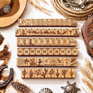 May include: Wooden game tiles with engraved Western-themed images and text. The tiles feature images of cowboys, boots, cacti, and directional letters. The word "JOKER" is visible on some tiles. Other Western accessories, including a hat, rope, and horseshoe, are also present.