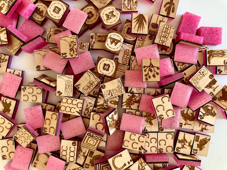 May include: A collection of wooden and pink resin game pieces. The wooden pieces feature etched designs of cacti, horseshoes, and cowboy boots. The pink resin pieces are rectangular and scattered among the wooden pieces.