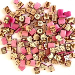 May include: A collection of small, wooden tiles with etched designs and pink, rectangular blocks. The wooden tiles feature various illustrations, including horseshoes, cacti, and other symbols. The pink blocks are scattered among the wooden tiles.