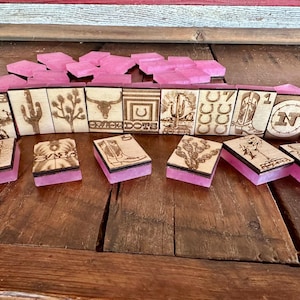 May include: A set of wooden dominoes with laser-etched western-themed designs, including a cactus, horseshoe, and bull skull. The dominoes have pink resin bases and are arranged on a wooden surface.
