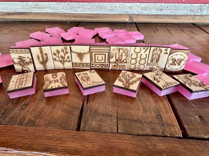 May include: A set of wooden dominoes with laser-etched western-themed designs, including a cactus, horseshoe, and bull skull. The dominoes have pink resin bases and are arranged on a wooden surface.