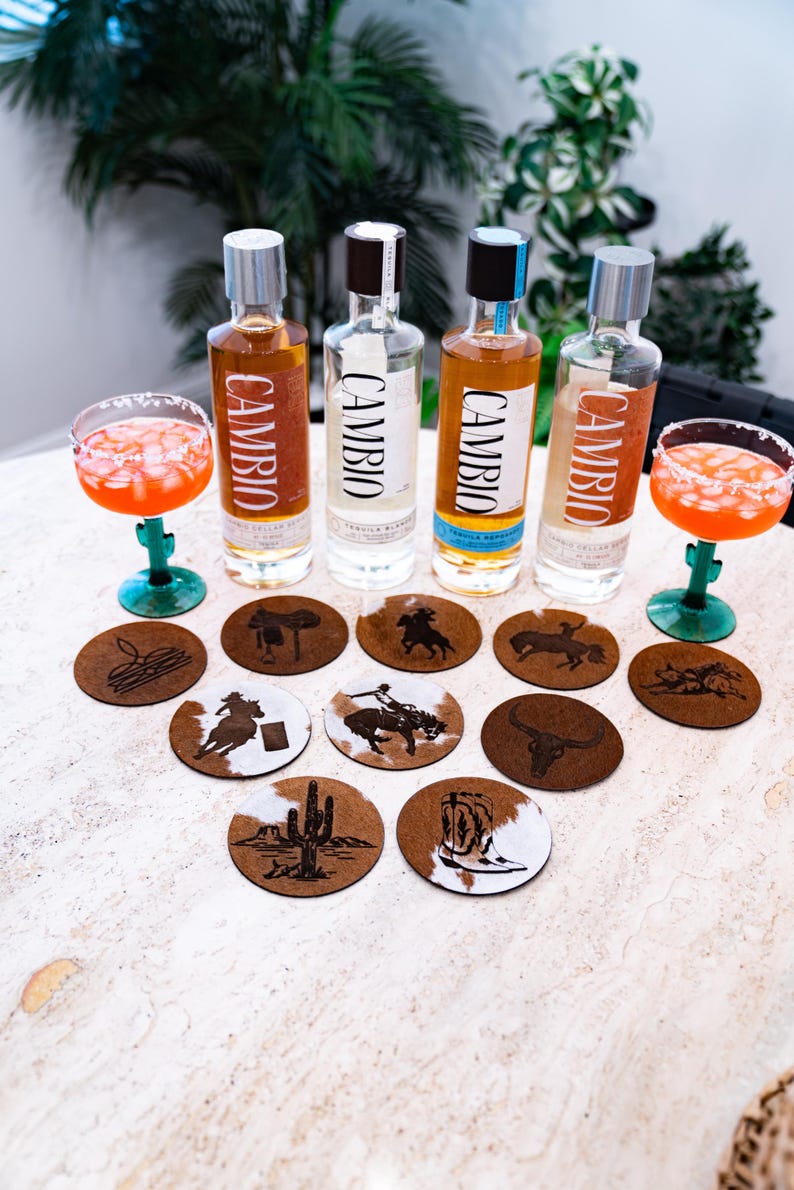 Peut inclure: Bouteilles de tequila avec le mot "CAMBIO" sur l'&eacute;tiquette, accompagn&eacute;es de cocktails dans des verres &agrave; pied verts. Des dessous de verre ronds avec des motifs de cow-boy sont dispos&eacute;s devant les bouteilles.