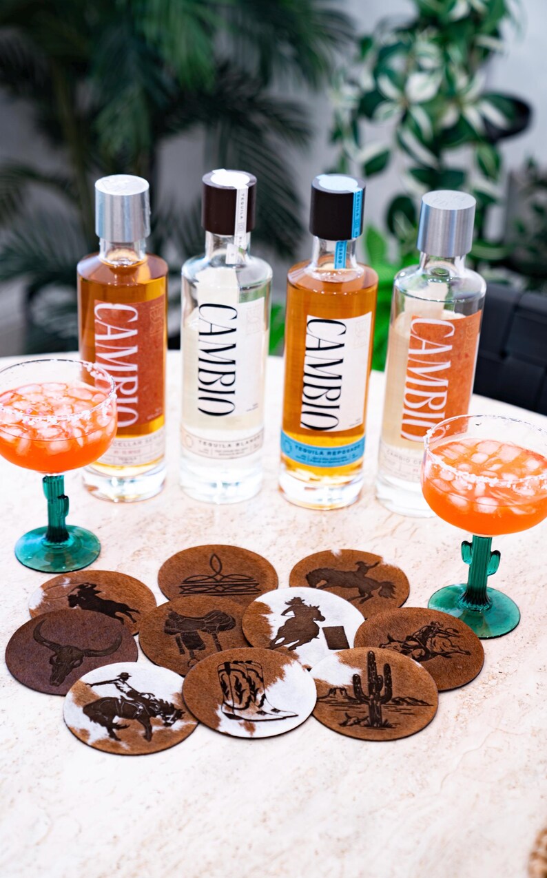 Peut inclure: Quatre bouteilles de tequila avec l'inscription "CAMBIO", accompagn&eacute;es de deux margaritas dans des verres &agrave; pied verts. Des dessous de verre ronds avec des motifs western sont &eacute;galement visibles.