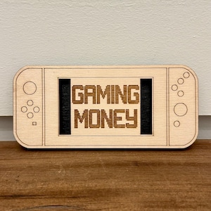 May include: A wooden gaming-themed decorative piece shaped like a game console. The text "GAMING MONEY" is displayed in pixelated font within a black rectangle. The piece is light brown with etched details.