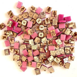 May include: A collection of wooden game pieces and pink resin squares scattered on a white surface. The wooden pieces feature engraved designs of cacti, horseshoes, and playing card suits. The pink squares are smooth and rectangular.