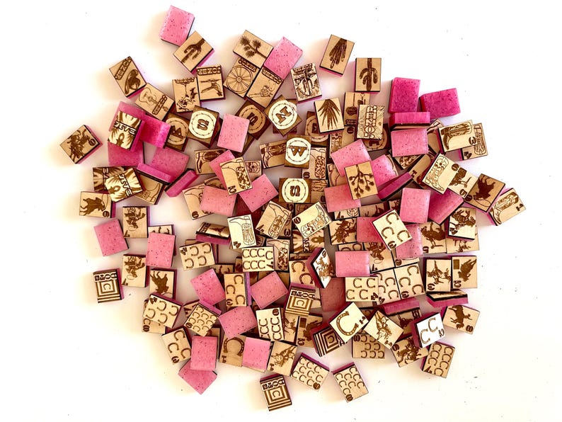 May include: A collection of wooden game pieces and pink resin squares scattered on a white surface. The wooden pieces feature engraved designs of cacti, horseshoes, and playing card suits. The pink squares are smooth and rectangular.