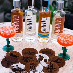Peut inclure: Quatre bouteilles de tequila Cambio avec diff&eacute;rentes &eacute;tiquettes et un cocktail margarita dans un verre vert en forme de cactus. Plusieurs dessous de verre ronds avec des motifs sur le th&egrave;me de l'Ouest sont &eacute;galement visibles.