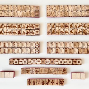 May include: Wooden game tiles with etched designs, including horseshoes, cowboys, cowboy boots, and cacti. The tiles are arranged in rows and include the letters "N", "W", "S", and "E". The tiles are displayed on a white surface.