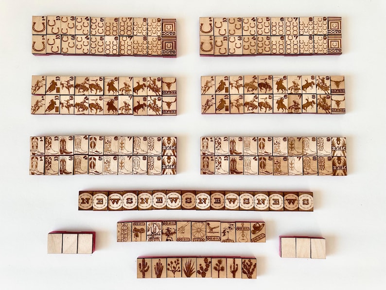 May include: Wooden game tiles with etched designs, including horseshoes, cowboys, cowboy boots, and cacti. The tiles are arranged in rows and include the letters "N", "W", "S", and "E". The tiles are displayed on a white surface.