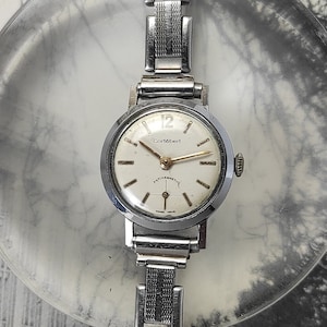 May include: A silver wristwatch with a white face and black hands. The watch has a silver metal band and says "Corbert" on the face.