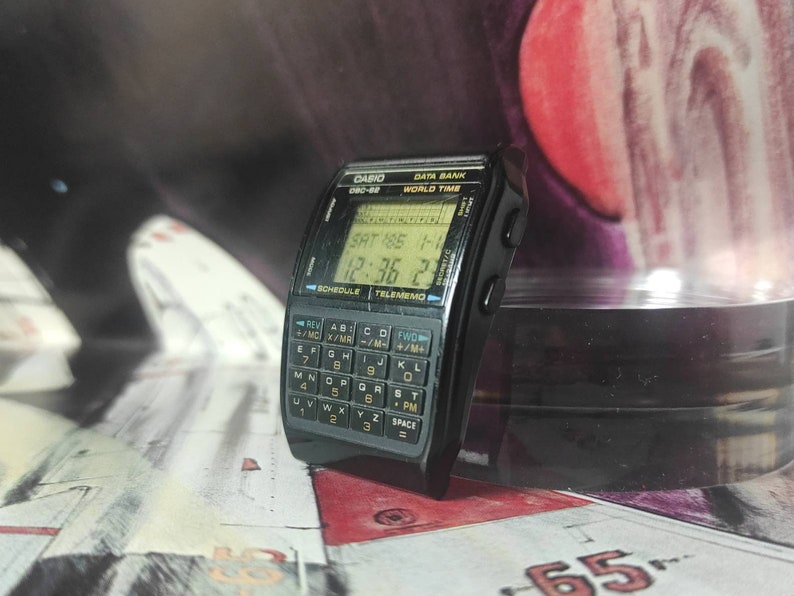 May include: A black Casio Data Bank watch with a digital display showing the date, time, and day of the week. The watch also has a calculator and a world time function. The watch is on a black background.
