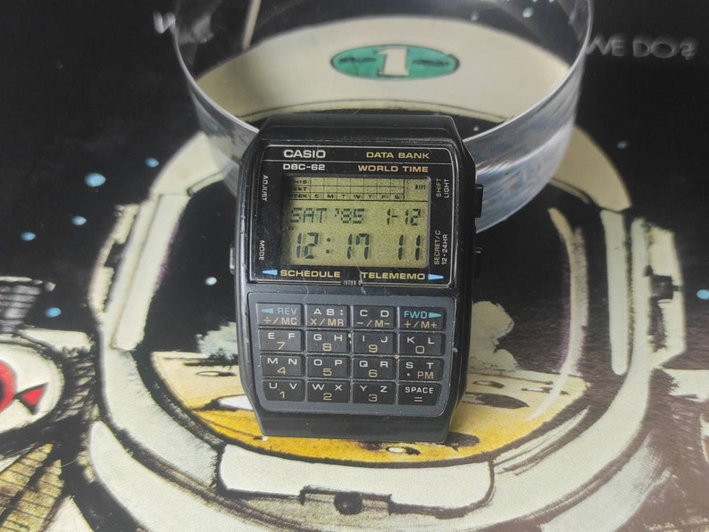 May include: A black Casio DBC-62 Data Bank watch with a digital display showing the time, date, and day of the week. The watch also has a calculator and a telememo function.