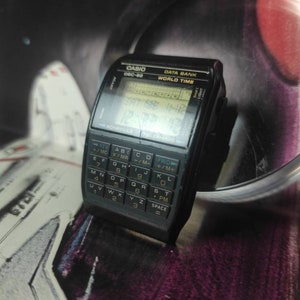 May include: A black Casio Data Bank World Time watch with a calculator face. The watch has a digital display showing the time and date. The calculator face has a QWERTY keyboard and a variety of functions.