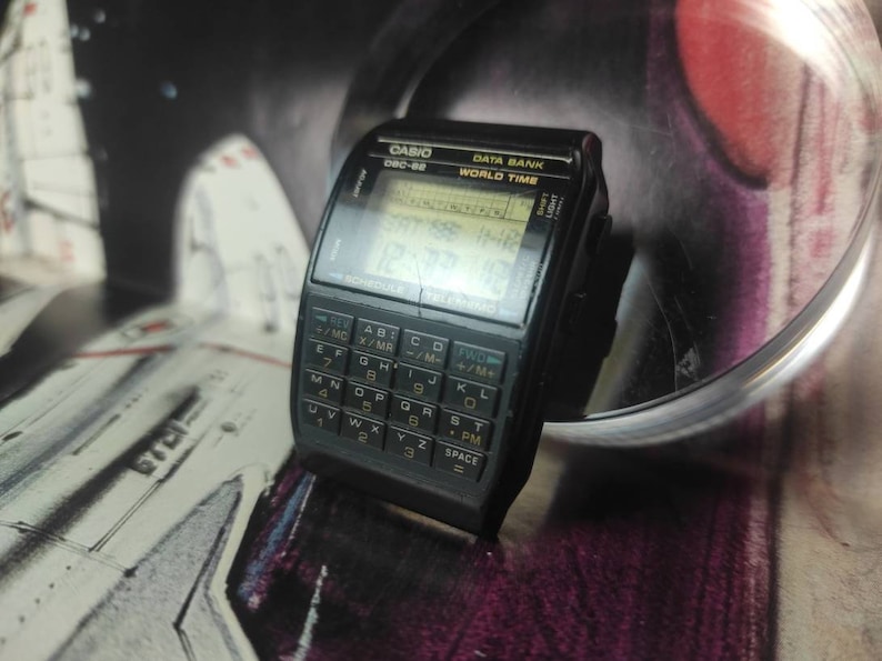 May include: A black Casio Data Bank World Time watch with a calculator face. The watch has a digital display showing the time and date. The calculator face has a QWERTY keyboard and a variety of functions.