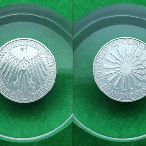 Può includere: Una moneta tedesca da 10 marchi tedeschi d'argento che commemora le Olimpiadi di Monaco di Baviera del 1972. Il dritto presenta l'aquila tedesca con il testo "Bundesrepublik Deutschland" e "10 Deutsche Mark". Il rovescio presenta un design stilizzato a raggi di sole con il testo "XX. Olympiade 1972 Spiele in München".