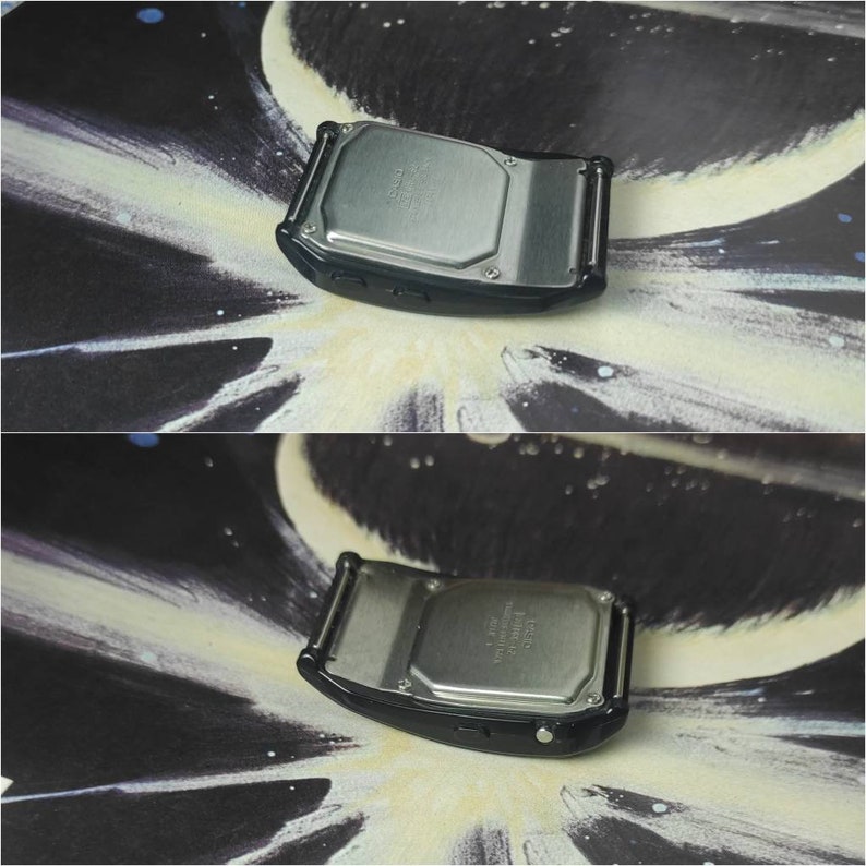 May include: The back of a black digital watch with a silver metal case. The watch has a silver metal band and a black button on the side.