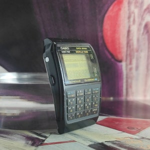 May include: A black Casio Data Bank watch with a digital display and a calculator keypad. The watch has a world time function and a schedule feature. The watch is on a table with a red and purple background.