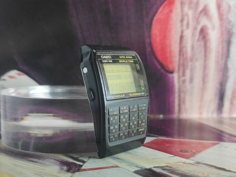 May include: A black Casio Data Bank watch with a digital display and a calculator keypad. The watch has a world time function and a schedule feature. The watch is on a table with a red and purple background.
