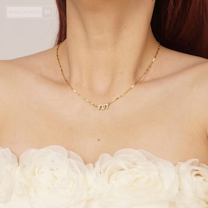 May include: A delicate gold necklace with a Hebrew inscription. The necklace features a thin chain and a pendant with the word in Hebrew. The necklace is worn against a white floral fabric.
