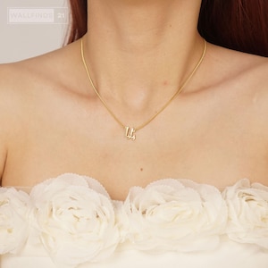 Könnte beinhalten: Eine goldfarbene Halskette mit einer zarten Kette und einem kleinen, stilisierten Anhänger mit arabischer Schrift. Der Anhänger ist mittig an der Kette platziert, die auf dem Schlüsselbein aufliegt. Das Design ist schlicht und glänzend.