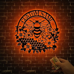 May include: A black metal bee and flower wreath with a customizable nameplate. The wreath is illuminated with warm orange light. A remote control is visible in the bottom right corner.