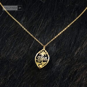 May include: A gold-toned necklace featuring a delicate chain and a pendant shaped like a seal. The pendant displays a cutout design of a palm tree, a sailboat, and the text GUAM, all enclosed within an oval border with a jagged edge.
