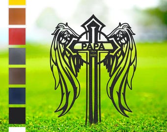 May include: A black metal memorial cross with angel wings, the word "PAPA" and the years 1944-2025. The cross is displayed on a green lawn, with a color palette of options on the left.
