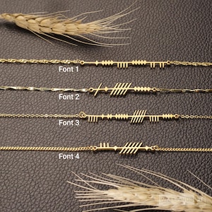 May include: Four gold-colored necklaces with unique bar and line designs, labeled Font 1 through Font 4. The necklaces are displayed on a dark brown surface, with wheat stalks in the background.
