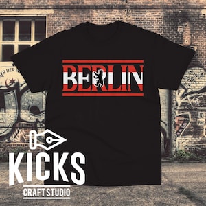 May include: Black t-shirt with the word "BERLIN" in white letters with red accents and a black bear graphic. The shirt also features the text "KICKS CRAFT STUDIO" in white.