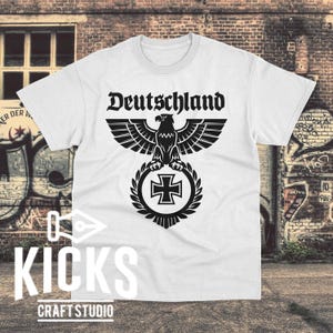 May include: White t-shirt featuring the word "Deutschland" above a black eagle emblem with a cross. The shirt has a crew neck and short sleeves. The bottom of the shirt has the text "KICKS CRAFT STUDIO".
